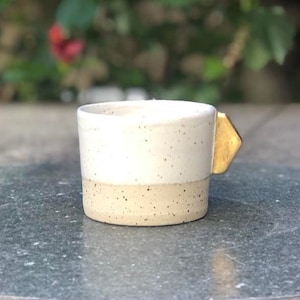 Peut inclure: Une petite tasse en céramique blanche avec un glaçage moucheté et une anse dorée. La tasse est posée sur une table grise.