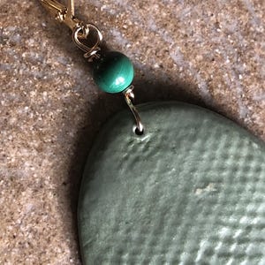 May include: Close-up of a single earring with a gold-colored leverback clasp. A small, round, green malachite bead is wire-wrapped above a textured, oval-shaped, sage green ceramic pendant. The earring is displayed against a neutral background.
