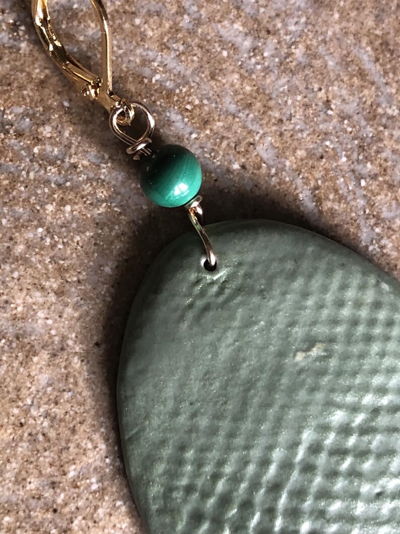 May include: Close-up of a single earring with a gold-colored leverback clasp. A small, round, green malachite bead is wire-wrapped above a textured, oval-shaped, sage green ceramic pendant. The earring is displayed against a neutral background.