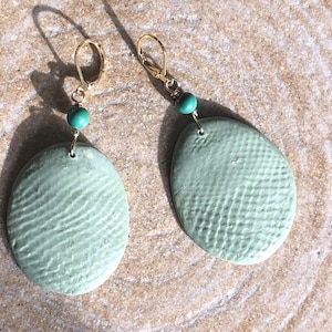 Peut inclure: Paire de boucles d'oreilles faites à la main avec des pendentifs ovales en céramique vert pâle texturé. Chaque boucle d'oreille a un crochet doré et une petite perle turquoise ronde. Présentées sur une surface texturée brun clair.