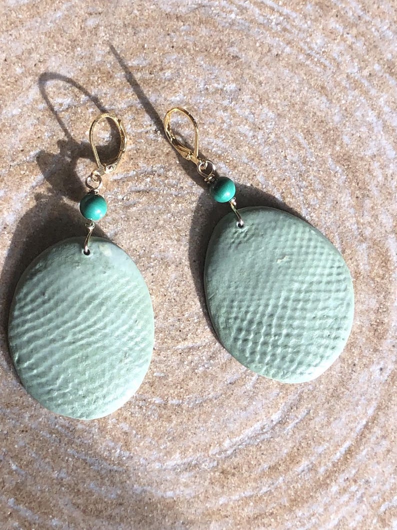 May include: A pair of handmade earrings featuring oval, textured, pale green ceramic pendants. Each earring has a gold-colored hook and a small, round, turquoise bead. The earrings are displayed on a light brown, textured surface.