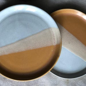 Puede incluir: Dos platos de cerámica con un diseño de bloques de color diagonal. Uno presenta una combinación de colores azul claro y amarillo mostaza, mientras que el otro tiene una combinación de colores amarillo mostaza y azul claro. Los platos son redondos y tienen un borde ligeramente elevado.