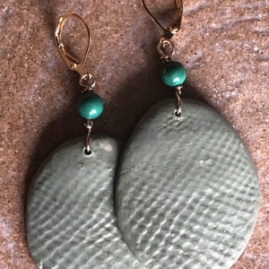 May include: A pair of handmade earrings featuring oval-shaped, textured, sage green pendants. Each earring has a gold-colored leverback clasp and a small, round, teal bead. The earrings are displayed on a textured, light brown surface.