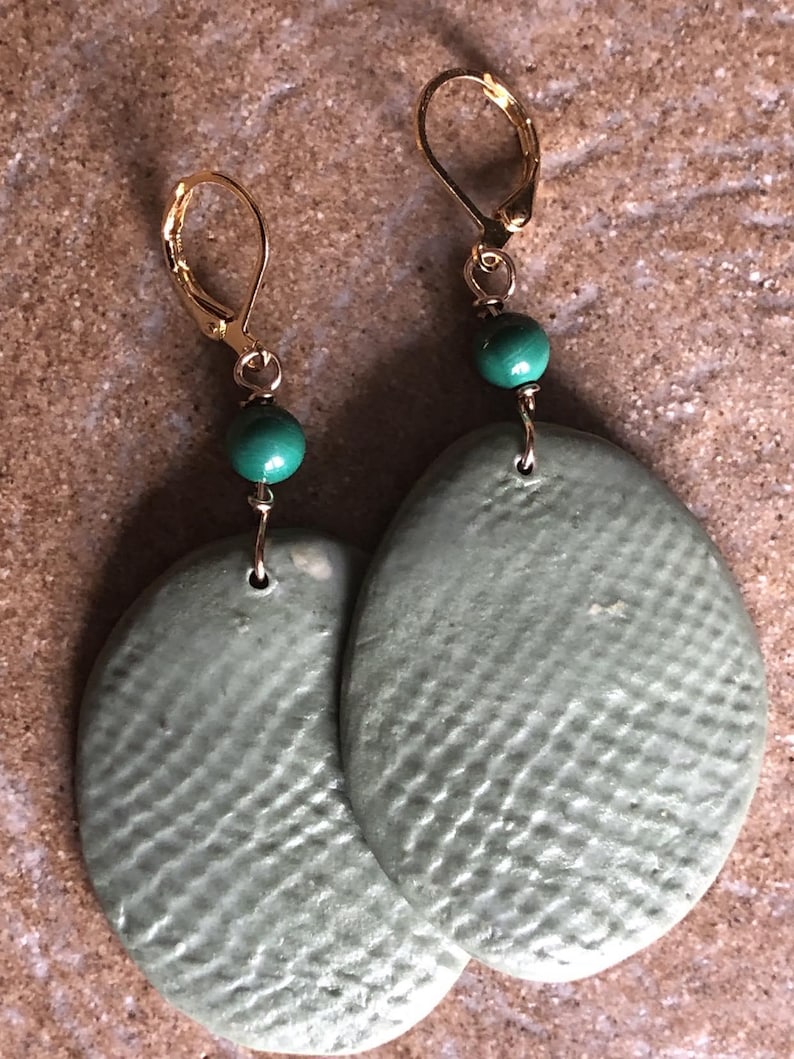 May include: A pair of handmade earrings featuring oval-shaped, textured, sage green pendants. Each earring has a gold-colored leverback clasp and a small, round, teal bead. The earrings are displayed on a textured, light brown surface.