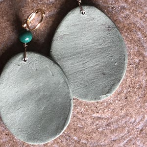 May include: Handmade earrings featuring large, oval-shaped, sage green pendants. Each earring has a gold-colored hoop and a small, round, green bead. The earrings are displayed on a textured, brown surface.