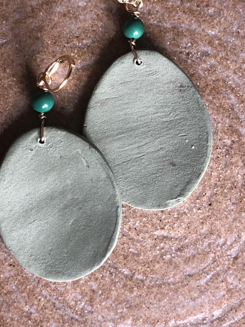 May include: Handmade earrings featuring large, oval-shaped, sage green pendants. Each earring has a gold-colored hoop and a small, round, green bead. The earrings are displayed on a textured, brown surface.