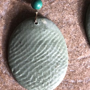 May include: Close-up of a pair of earrings. Each earring features a textured, oval-shaped, pale green pendant suspended from a gold-colored wire. A small, round, teal bead sits above each pendant, adding a pop of color.