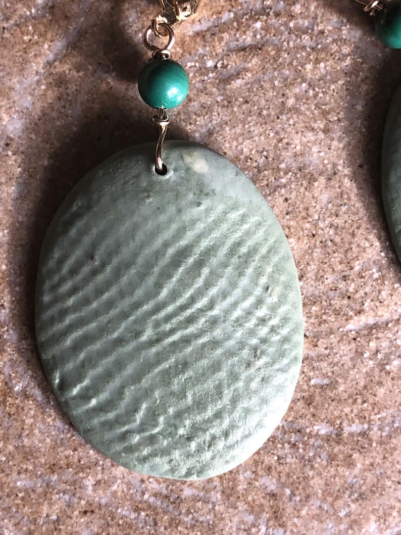 May include: Close-up of a pair of earrings. Each earring features a textured, oval-shaped, pale green pendant suspended from a gold-colored wire. A small, round, teal bead sits above each pendant, adding a pop of color.