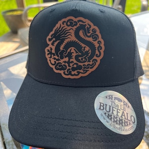 May include: Black trucker hat with a brown embroidered dragon design. The hat has a black mesh back and a black brim. The hat has a sticker on the front that says "The Lil Buffalo" and a website address.