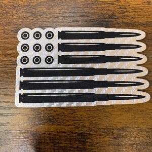 May include: A sticker in the shape of a flag, featuring a design of bullets in place of stripes and circles for stars. The bullets are black, and the background is silver. The sticker is on a brown wooden surface.