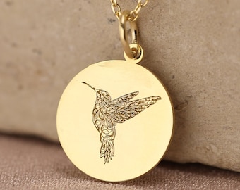 Puede incluir: Un collar con colgante circular de tono dorado que presenta un diseño delicado de un colibrí grabado en el centro. El colgante cuelga de una cadena fina a juego del mismo tono, mostrando un acabado metálico pulido y una forma redonda.