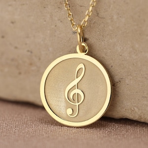 Op de afbeelding: Een gouden ketting met een ronde hanger met een mat afgewerkt midden en een gepolijste gouden rand. De hanger is in het midden gegraveerd met een vioolsleutel en hangt aan een fijne gouden ketting met kleine, uniforme schakels.
