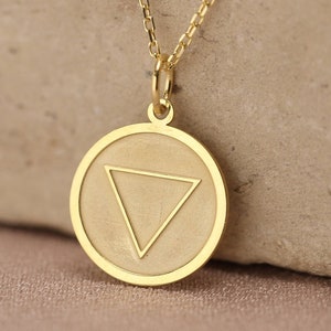 May include: A gold necklace with a round pendant featuring a triangle symbol. The pendant is on a chain and is resting on a light brown surface.