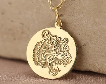 May include: A gold-colored pendant necklace featuring a detailed tiger head design within a circular pendant. The necklace has a delicate chain and is displayed on a neutral-toned surface. The pendant is approximately 1 inch in diameter.