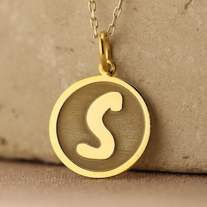 Puede incluir: Un collar de oro con un colgante redondo que presenta la letra 'S' en una fuente cursiva.