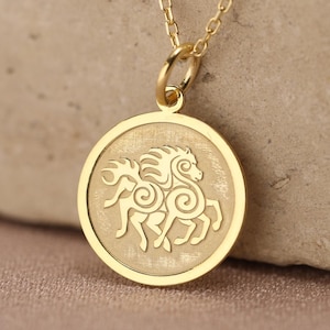 Può includere: Una collana color oro con un ciondolo circolare caratterizzato da un bordo lucido e un centro opaco. Il ciondolo presenta l'incisione di un cavallo stilizzato con motivi a spirale sul corpo ed è appeso a una sottile catenina dorata.