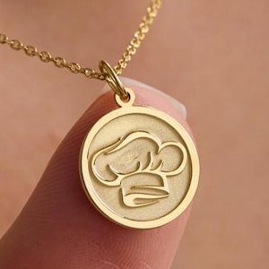 May include: A gold-toned circular pendant necklace featuring a central engraved chef hat design. The pendant is suspended from a delicate gold-toned chain. The circular charm has a smooth, polished border surrounding a matte-textured interior surface.
