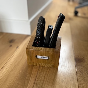 May include: A wooden box with the word "REMOTES" on a small metal label, holding several black remote controls. The box is made of light-colored wood and sits on a wooden floor.