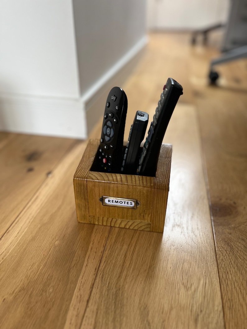 May include: A wooden box with the word "REMOTES" on a small metal label, holding several black remote controls. The box is made of light-colored wood and sits on a wooden floor.