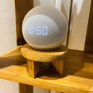Puede incluir: Un altavoz inteligente blanco con un reloj digital que muestra las 8:50 en un soporte de madera con tres patas.