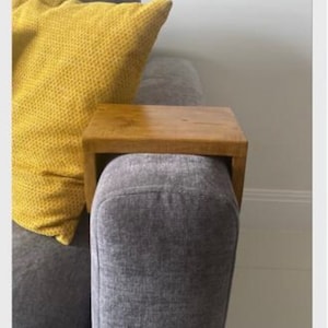 May include: A wooden armrest tray for a sofa or armchair. The tray is made of natural wood and has a rustic finish. It is placed on a gray sofa with a yellow throw pillow.