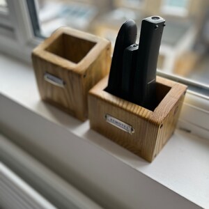 May include: Two wooden remote control holders sit on a windowsill. One holder is empty, the other contains three black remotes. Both holders are labeled with small metal tags. The holders are made of light-colored wood.