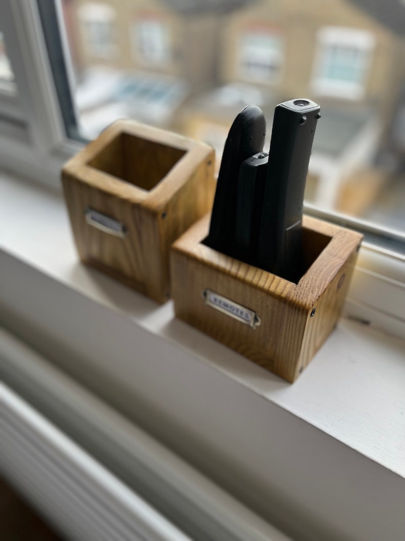 May include: Two wooden remote control holders sit on a windowsill. One holder is empty, the other contains three black remotes. Both holders are labeled with small metal tags. The holders are made of light-colored wood.