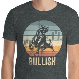 May include: A dark grey t-shirt featuring a vintage-style graphic of a black bull charging against a colourful sunset background. The graphic includes a stock market chart and the word "BULLISH" in white text.