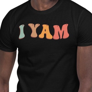 以下が含まれることがあります： 緑、オレンジ、赤の色でレトロなスタイルの文字で「I YAM」と書かれた黒いTシャツです。