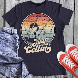 May include: A navy blue t-shirt with a vintage-style graphic of a cello and musical notes. The text "Just Cellin'" is printed in a retro font below the graphic.