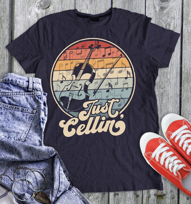 May include: A navy blue t-shirt with a vintage-style graphic of a cello and musical notes. The text "Just Cellin'" is printed in a retro font below the graphic.