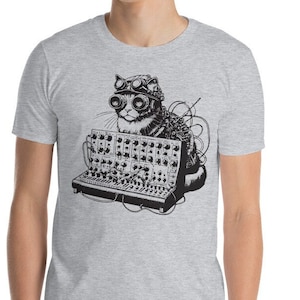 Può includere: T-shirt grigio chiaro con un'illustrazione in bianco e nero di un gatto con occhiali e un complesso dispositivo elettronico. Il gatto è al centro, con linee e ombreggiatura dettagliate. Sono visibili fili e manopole.