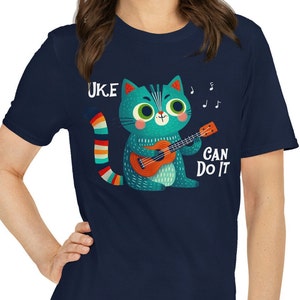 May include: A navy blue t-shirt with a cartoon cat playing a ukulele. The cat is blue and green with a rainbow tail. The text "UKE CAN DO IT" is printed on the shirt.