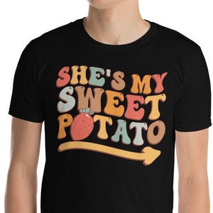 以下が含まれることがあります： 「She's My Sweet Potato」というレトロなデザインのグラフィックがプリントされた黒いTシャツ。漫画のさつまいもが描かれている。