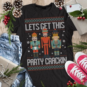 May include: Black t-shirt with a Christmas-themed design featuring three nutcracker figures and the text "Let's Get This Party Crackin'" in a white, red, and green colour scheme.