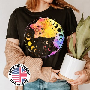 Könnte beinhalten: Schwarzes T-Shirt mit einem bunten Grafikdruck von zwei Katzen in einem Yin-Yang-Symbol mit Monden und Sternen. Die Grafik ist im Aquarellstil mit rosa, orange, gelben und blauen Farben gehalten. Der Text "FREE DELIVERY USA and UK" befindet sich auf einem weißen Kreis mit einer rot-weiß-blauen Flagge.
