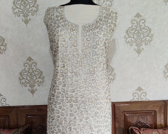 May include: A cream-coloured sleeveless tunic with a V-neckline and a silver and brown geometric pattern. The tunic is displayed on a mannequin against a patterned wallpaper background.
