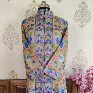 May include: A long, beige embroidered coat with a mandarin collar and floral patterns in shades of blue, purple, and green. The coat has a button closure and long sleeves.