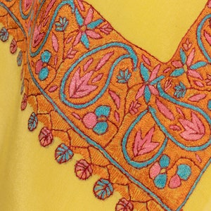 May include: Close-up of a yellow garment with intricate embroidery. The embroidery features a paisley and floral design in orange, pink, and teal. The detailed stitching creates a decorative border along the neckline, showcasing a blend of colors and patterns.