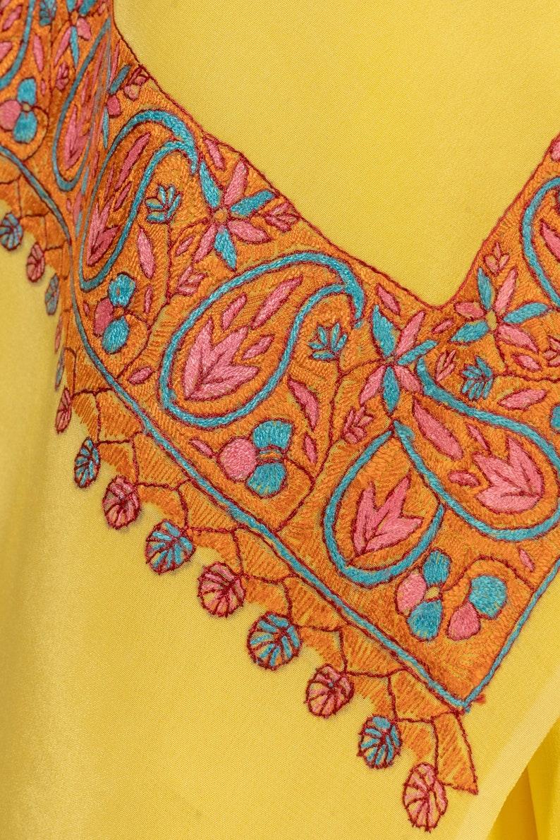 May include: Close-up of a yellow garment with intricate embroidery. The embroidery features a paisley and floral design in orange, pink, and teal. The detailed stitching creates a decorative border along the neckline, showcasing a blend of colors and patterns.
