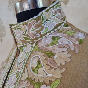 K&ouml;nnte beinhalten: Eine braune Jacke mit einem hohen Kragen, der mit aufwendigen Stickereien in Wei&szlig;-, Gr&uuml;n- und Beiget&ouml;nen verziert ist. Die Stickerei zeigt ein Blumenmuster mit Paisley-Motiven.