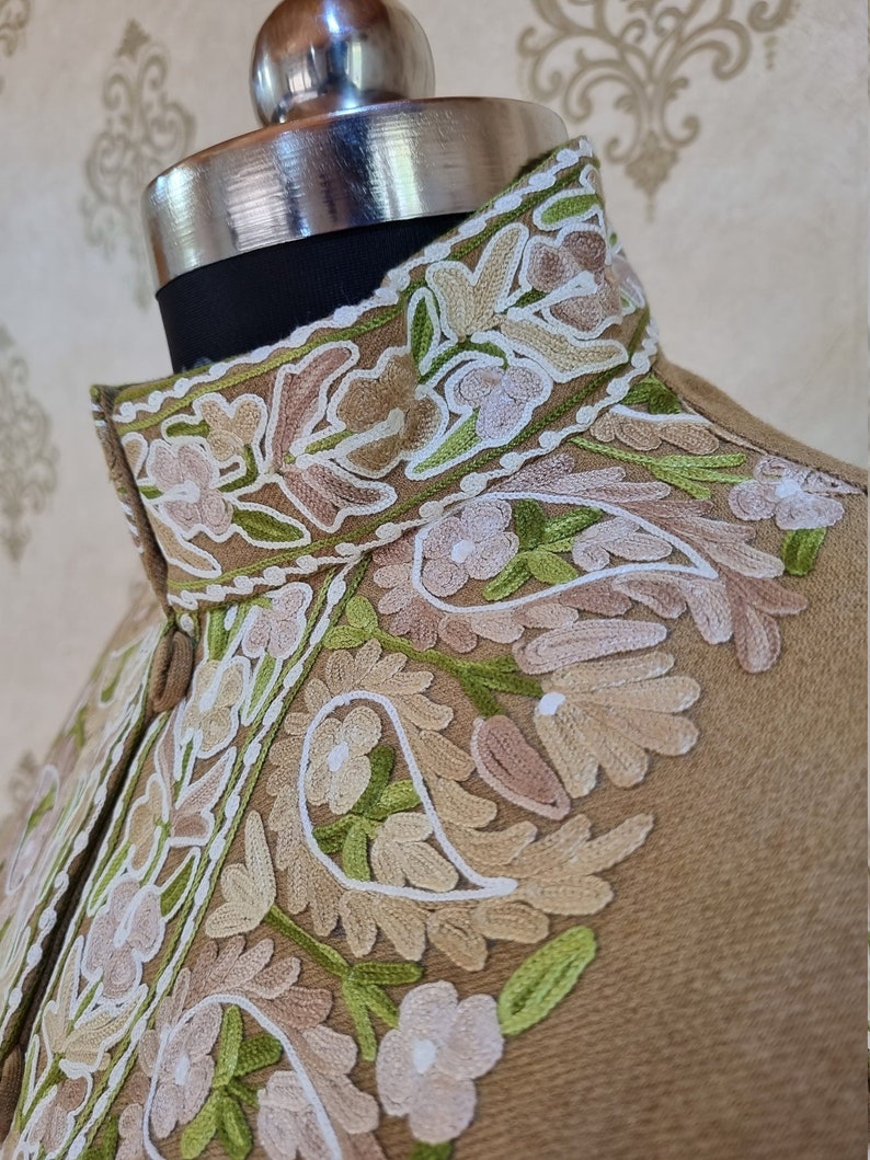 K&ouml;nnte beinhalten: Eine braune Jacke mit einem hohen Kragen, der mit aufwendigen Stickereien in Wei&szlig;-, Gr&uuml;n- und Beiget&ouml;nen verziert ist. Die Stickerei zeigt ein Blumenmuster mit Paisley-Motiven.