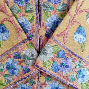 May include: A tan jacket with intricate floral embroidery in shades of blue, green, and pink. The embroidery features a variety of flower shapes and sizes, creating a delicate and feminine design.