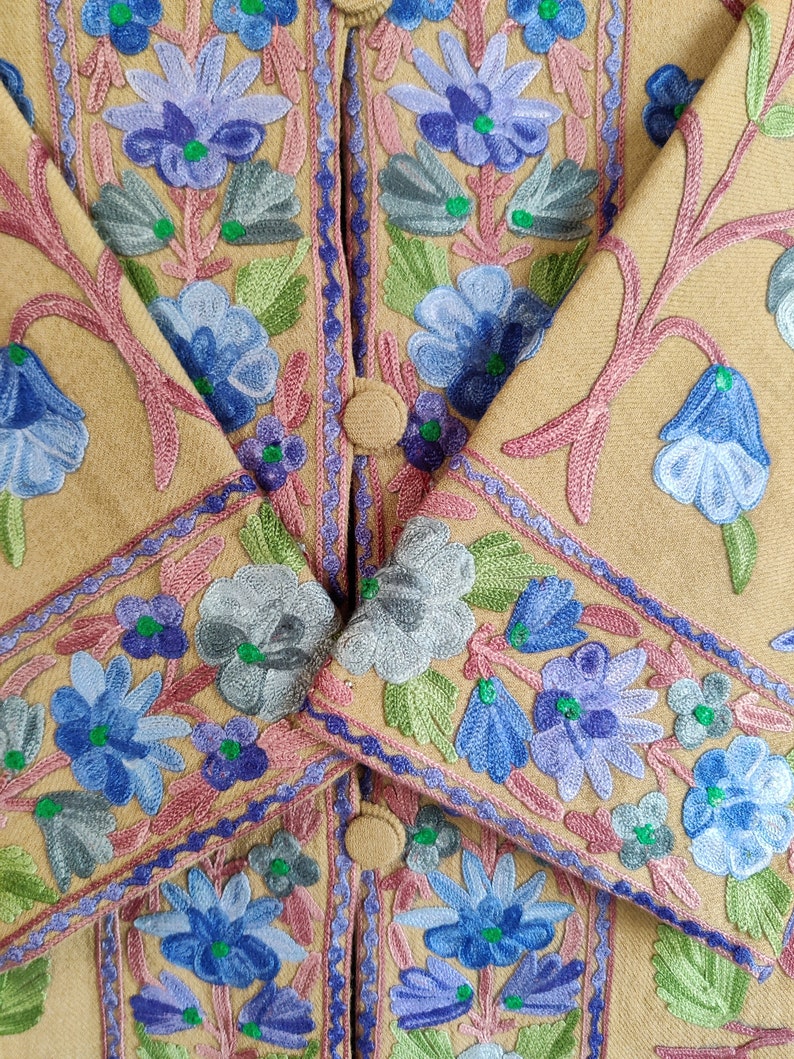 May include: A tan jacket with intricate floral embroidery in shades of blue, green, and pink. The embroidery features a variety of flower shapes and sizes, creating a delicate and feminine design.