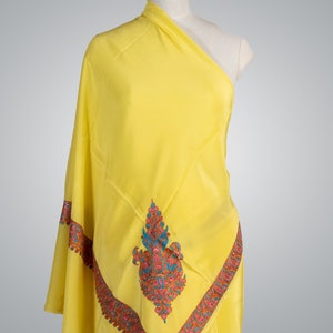 May include: A bright yellow shawl draped over a mannequin. The shawl features an intricate embroidered border and central design in shades of pink, blue, and orange. The fabric appears soft and flowing, suggesting a luxurious textile.