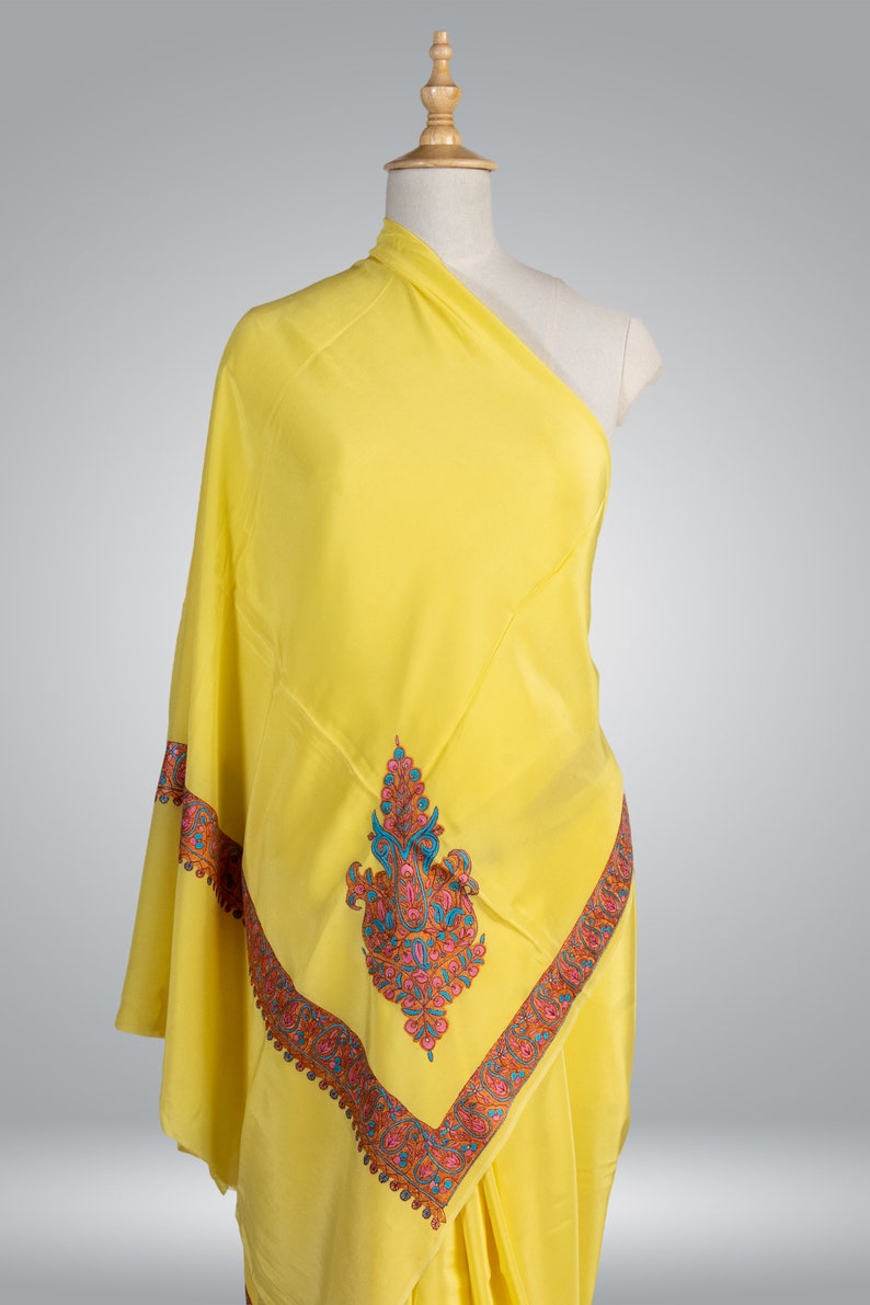 May include: A bright yellow shawl draped over a mannequin. The shawl features an intricate embroidered border and central design in shades of pink, blue, and orange. The fabric appears soft and flowing, suggesting a luxurious textile.