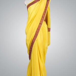 May include: A vibrant yellow saree draped on a mannequin. The saree features a decorative border with red, green, and blue floral patterns. The fabric appears smooth and flowing, showcasing the traditional Indian garment's elegant design.
