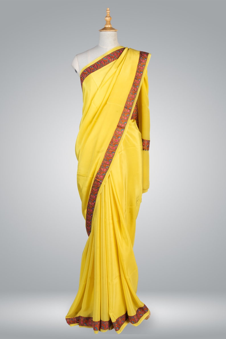 May include: A vibrant yellow saree draped on a mannequin. The saree features a decorative border with red, green, and blue floral patterns. The fabric appears smooth and flowing, showcasing the traditional Indian garment's elegant design.