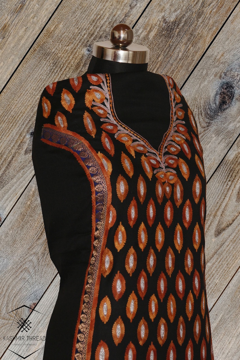 May include: Black fabric with intricate embroidery in orange, silver, and blue. The embroidery features a repeating pattern of leaves and a floral design around the neckline.