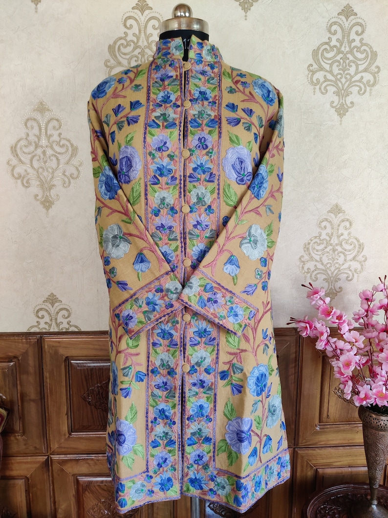 May include: A long, beige embroidered coat with a mandarin collar and floral patterns in shades of blue, purple, and green. The coat has a button closure and long sleeves.
